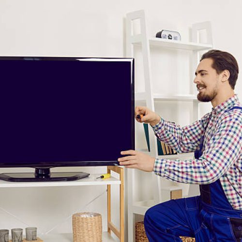 Repairman or technician fixing a TV at home. Young man in blue uniform overalls using a screwdriver while repairing, installing or checking a modern large television set in the living room