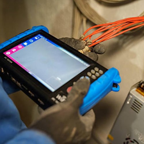 Close-up of a technician using an IPC tester to check BNC connectors and network cables during a security camera installation or maintenance task. The professional is wearing gloves and holding multiple coaxial connectors, with visible cabling and a power supply unit in the background. The image represents modern surveillance system setup, network diagnostics, and technical maintenance work in the field of security systems and electronics.